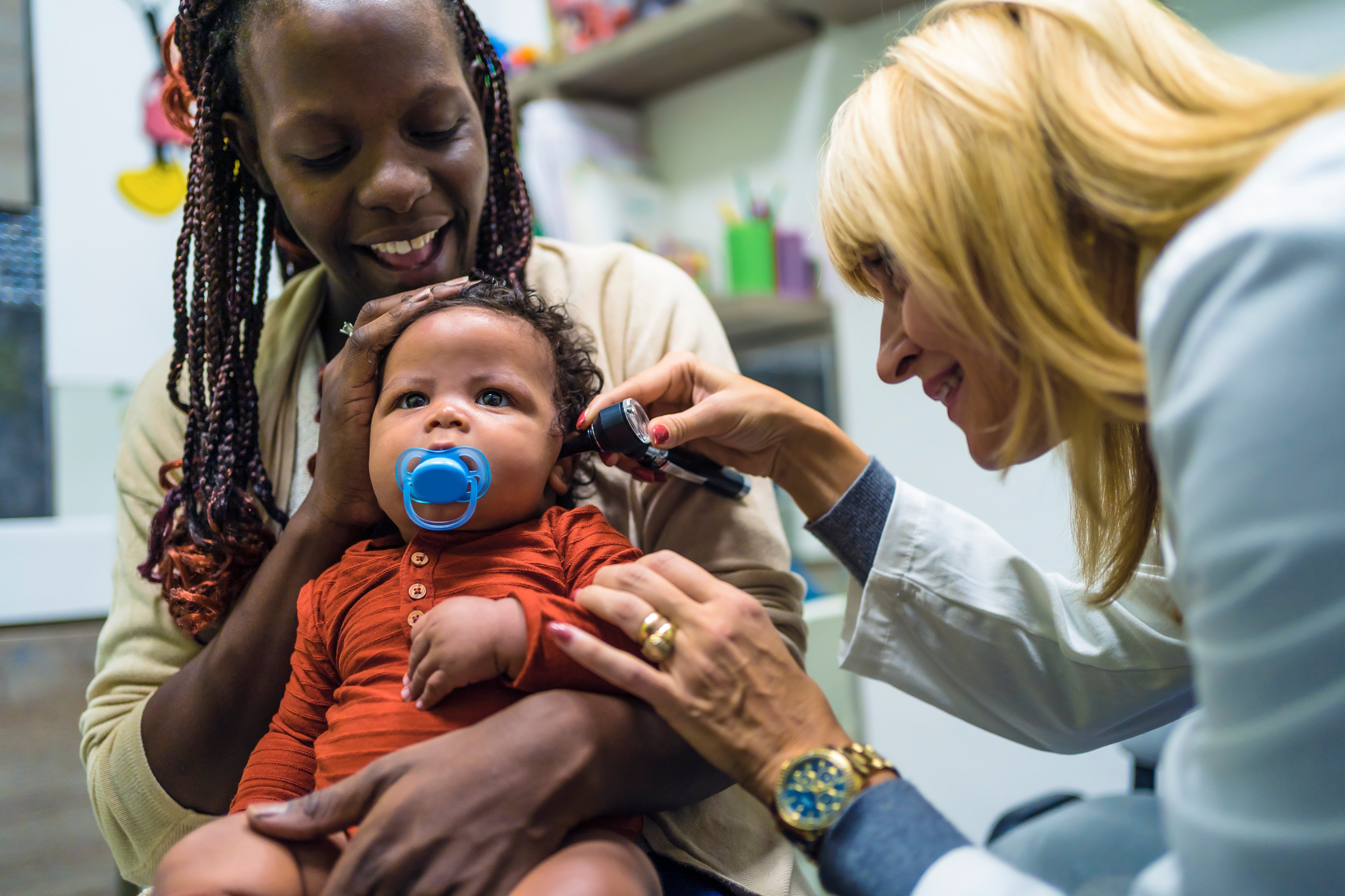 A doctor examines a baby's ear as his mother looks on, smiling