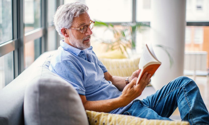 Older man in blue shirt reading a book on a couch