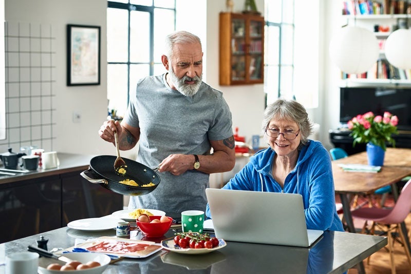 Senior woman using laptop and smiling as man serves breakfast
