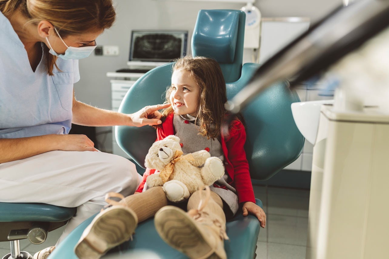 Dentist in protective face mask looking at girl sitting with stuffed toy